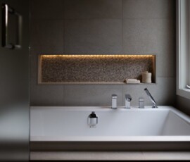 Luxurious bathtub with built-in illuminated niche for bath essentials, set against a modern tiled wall with a large window, highlighting sophisticated bathroom design services in Victoria, BC by Campo Design.