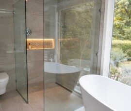 Modern bathroom with glass-enclosed shower, freestanding bathtub, and large window overlooking a garden, showcasing luxury bathroom renovation and design services in Victoria, BC by Campo Design.